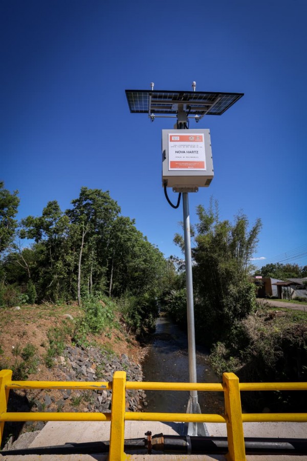 Close da estação de monitoramento instalada sobre ponte em Nova Hartz. O equipamento, identificado por uma placa do Cemaden e do Governo do Estado, coleta dados de chuva e nível de rios para prevenção de desastres. O cenário ao fundo inclui vegetação e o curso d’água monitorado.