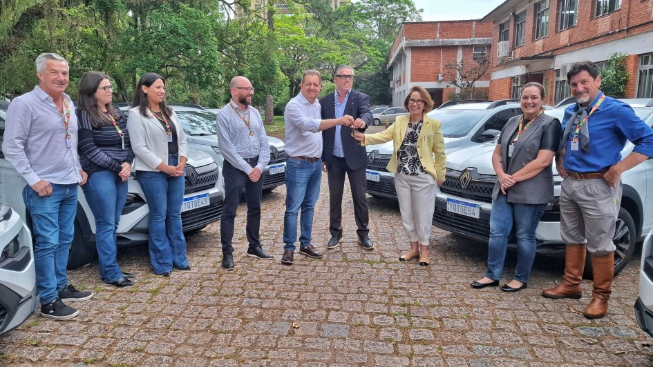 Nove pessoas estão em pé, posando para a foto, ao ar livre, com diversos carros estacionados ao fundo. No centro da imagem, dois homens e uma mulher seguram as chaves de um carro.