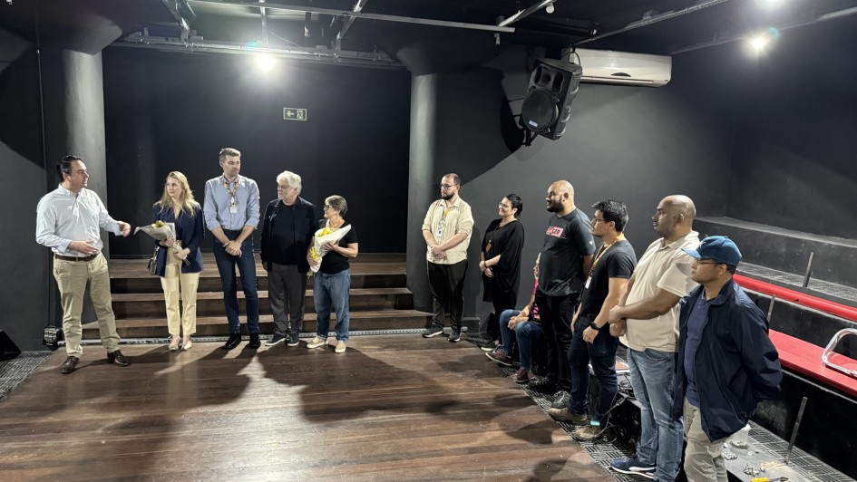 Foto de um palco de teatro, pequeno e rodeado de algumas poucas fileiras de arquibancada. Há 12 pessoas de pé no palco, em roda.