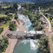 Uma ponte está sendo construída sobre um rio em uma área rural. Há estruturas metálicas, máquinas e materiais nos dois lados do rio. Casas e vegetação cercam o local, e a obra conecta duas partes da comunidade.