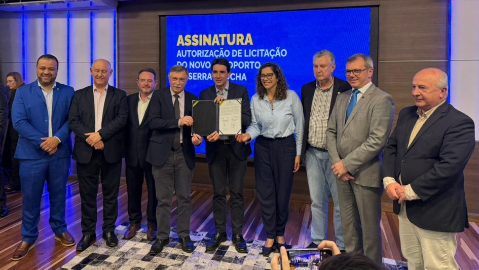 Na imagem, um grupo de nove pessoas posa para foto durante cerimônia de assinatura de autorização da licitação do novo Aeroporto da Serra Gaúcha. Ao centro, dois homens seguram o documento assinado. Atrás deles, um telão exibe o texto “Assinatura – Autorização de Licitação do Novo Aeroporto da Serra Gaúcha”. O ambiente é fechado, com iluminação artificial e piso de madeira.