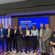 Na imagem, um grupo de nove pessoas posa para foto durante cerimônia de assinatura de autorização da licitação do novo Aeroporto da Serra Gaúcha. Ao centro, dois homens seguram o documento assinado. Atrás deles, um telão exibe o texto “Assinatura – Autorização de Licitação do Novo Aeroporto da Serra Gaúcha”. O ambiente é fechado, com iluminação artificial e piso de madeira.