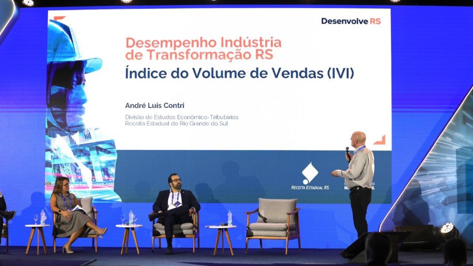A imagem mostra uma apresentação em evento sobre o desempenho da indústria de transformação do Rio Grande do Sul. No palco, um homem em pé fala ao microfone diante de um telão que exibe o título “Desempenho Indústria de Transformação RS – Índice do Volume de Vendas (IVI)”, com o nome do palestrante, André Luis Contri, da Receita Estadual. Sentados à esquerda, dois participantes acompanham a exposição. O ambiente é iluminado, com cenário azul e logotipo da Receita Estadual do RS projetado no painel.