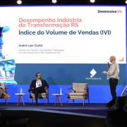A imagem mostra uma apresentação em evento sobre o desempenho da indústria de transformação do Rio Grande do Sul. No palco, um homem em pé fala ao microfone diante de um telão que exibe o título “Desempenho Indústria de Transformação RS – Índice do Volume de Vendas (IVI)”, com o nome do palestrante, André Luis Contri, da Receita Estadual. Sentados à esquerda, dois participantes acompanham a exposição. O ambiente é iluminado, com cenário azul e logotipo da Receita Estadual do RS projetado no painel.