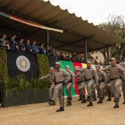 Governador Eduardo Leite participa da formatura de novos soldados da Brigada Militar. Na foto, ele está no palanque oficial com outras autoridades. Em primeiro plano, uma tropa de policiais militares passa marchando.
