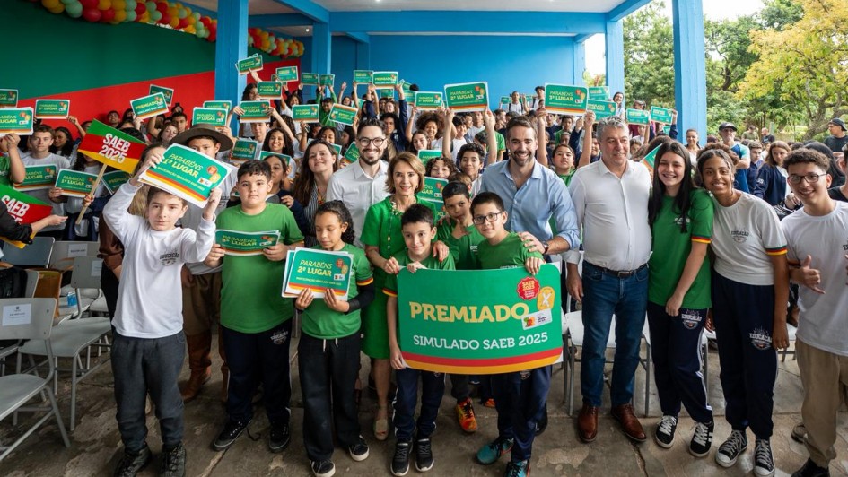 A imagem retrata um grupo numeroso de estudantes e adultos reunidos para uma foto coletiva, com o governador ao centro. Todos estão posicionados sob uma estrutura azul, segurando cartazes verdes com frases como “Premiado Simulado Saeb 2025”.