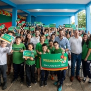 A imagem retrata um grupo numeroso de estudantes e adultos reunidos para uma foto coletiva, com o governador ao centro. Todos estão posicionados sob uma estrutura azul, segurando cartazes verdes com frases como “Premiado Simulado Saeb 2025”.