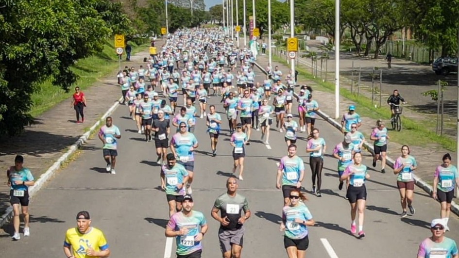 Grupo de corredores participa de prova de rua na avenida larga, com camisetas coloridas e números no peito. Clima de movimento, esporte e interação comunitária.