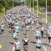 Grupo de corredores participa de prova de rua na avenida larga, com camisetas coloridas e números no peito. Clima de movimento, esporte e interação comunitária.