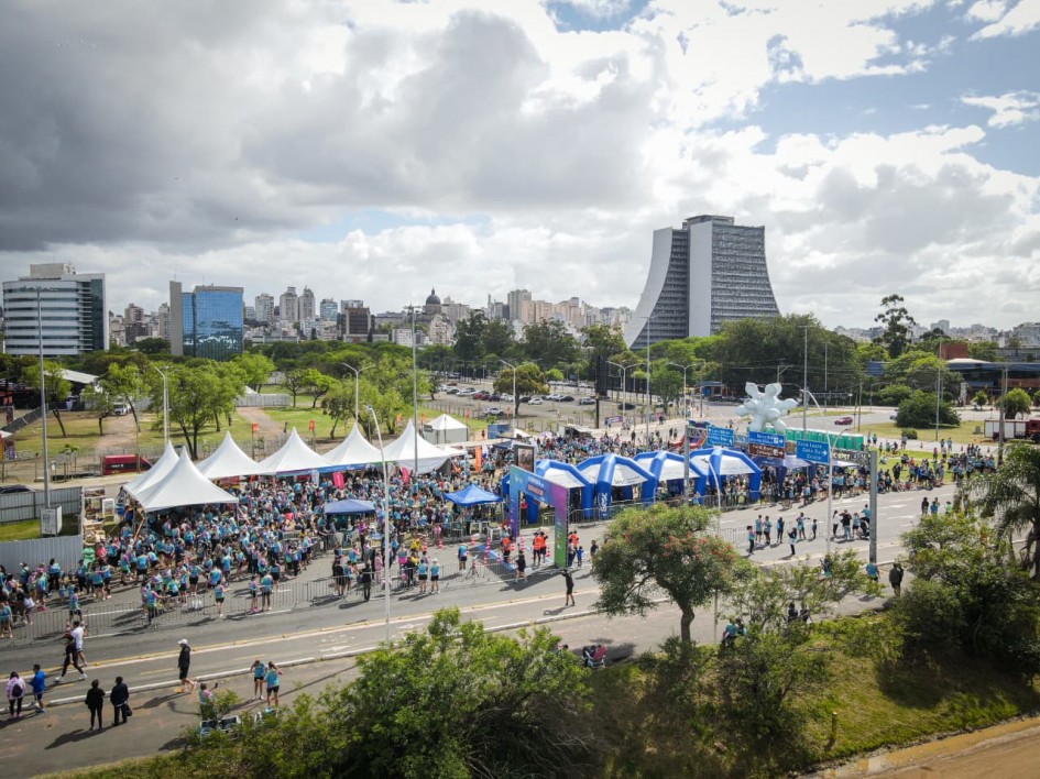 Evento ao ar livre em área urbana, com muitas pessoas reunidas perto de tendas brancas e azuis. Alguns caminham ou correm, e prédios modernos compõem o cenário. Clima festivo e bem organizado.