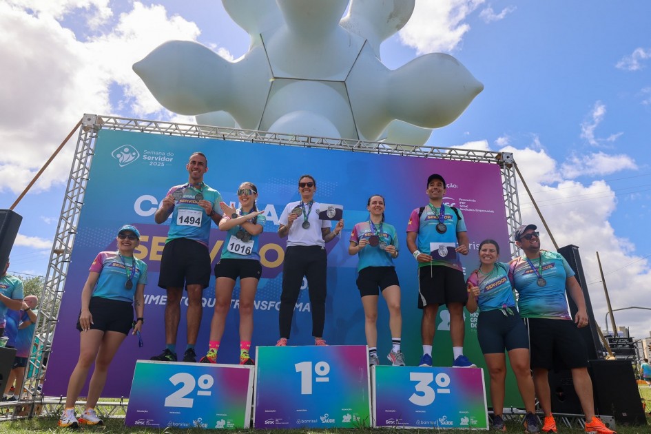 Três atletas em pódio comemoram vitória em corrida. Usei medalhas e camisetas esportivas com números. Ao fundo, painel colorido destaca nome do evento e mensagem de incentivo.