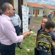 Homem entrega chave a mulher com criança no colo, em frente a casas novas. Cena transmite emoção e conquista, possivelmente entrega de moradia em evento comunitário.