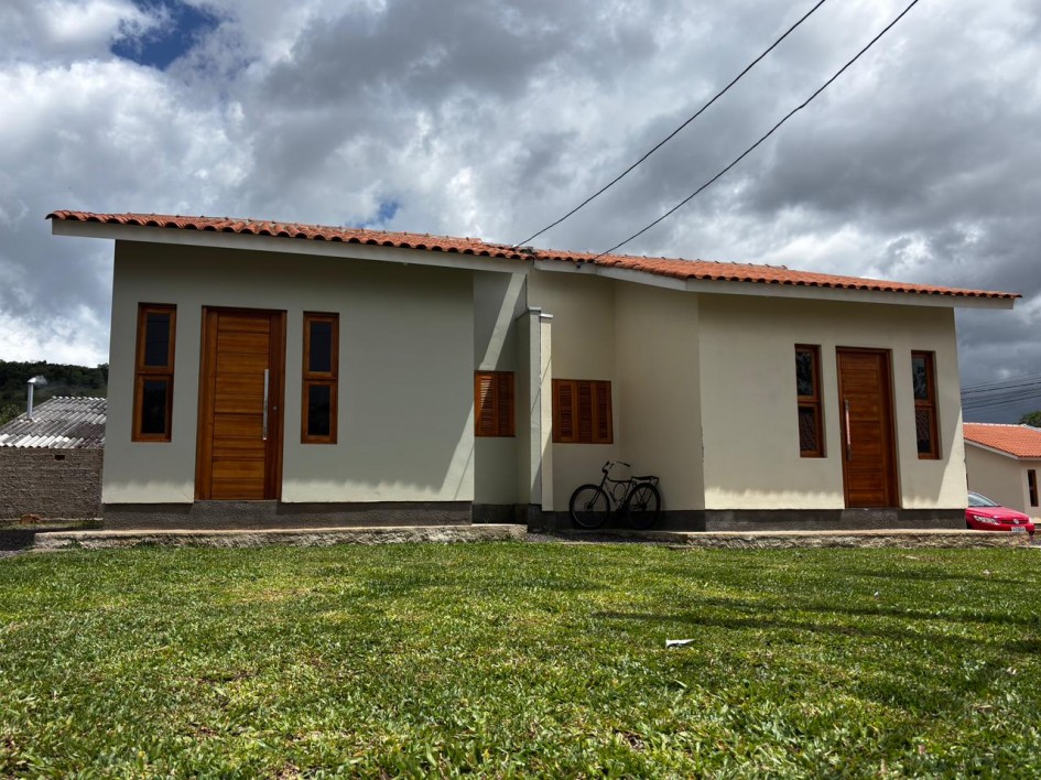 Casa térrea com telhado de cerâmica, duas entradas e janelas de madeira. Bicicleta encostada na parede e gramado ao redor. Ambiente simples e residencial.