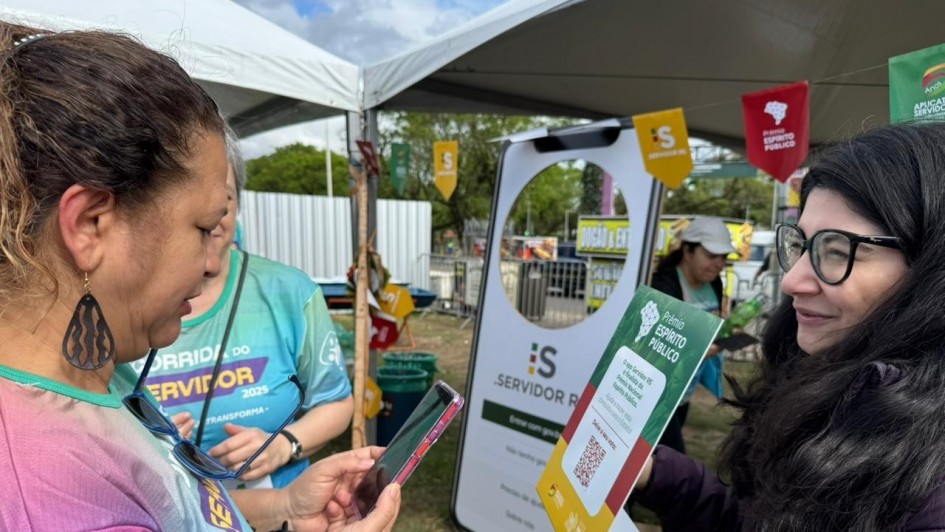 Duas pessoas interagem em estande de evento ao ar livre. Uma segurança celular, outra exibe cartaz com QR code e frase “Servidor Público”. Ambiente com tendas e clima de campanha.