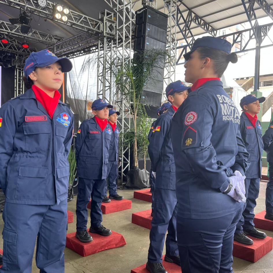 A imagem mostra algumas crianças em forma, perfiladas, uniformizadas em tom azul marinho e vermelho. No lado direito, há uma bombeiro militar olhando para a turma de crianças.
