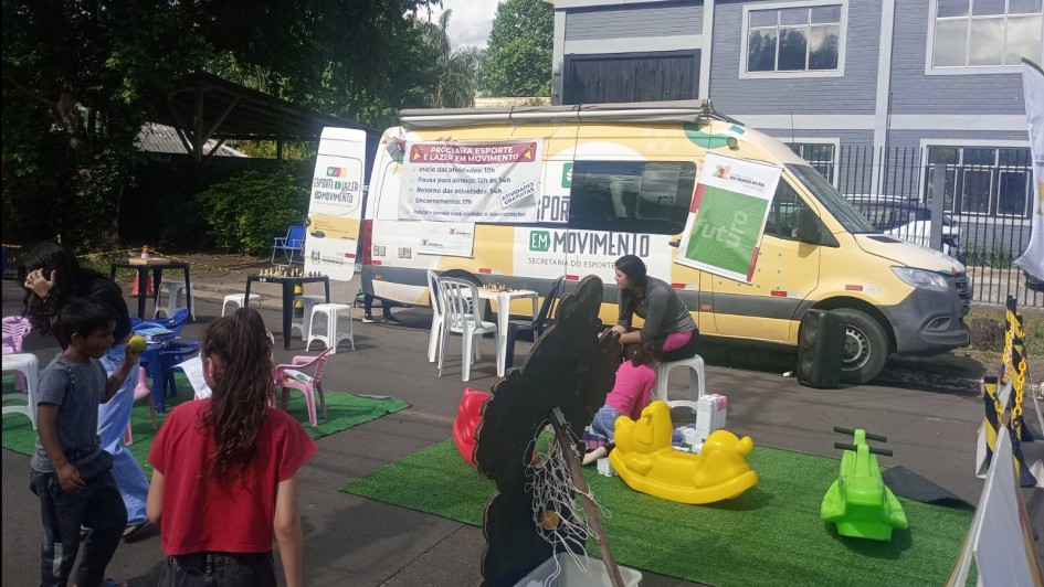 Foto mostra a van do Programa Esporte e Lazer em Movimento ao fundo, um furgão amarelo, com banners coloridos. Na frente, está montando brinquedos infantis. Uma mulher adulta acompanha várias crianças que circulam pelos brinquedos.