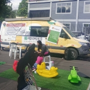 Foto mostra a van do Programa Esporte e Lazer em Movimento ao fundo, um furgão amarelo, com banners coloridos. Na frente, está montando brinquedos infantis. Uma mulher adulta acompanha várias crianças que circulam pelos brinquedos. 