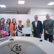 Foto posada em ambiente fechado de cor branca. Cinco mulheres e dois homens aparecem de pé, de frente para a câmera, em trajes formais, sorrindo. No meio, Arita segura e exibe o documento assinado.