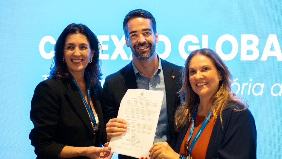 Uma foto de três pessoas (duas mulheres e um homem, no centro) em um evento, posando e segurando um documento (possivelmente um termo de cooperação ou parceria). Eles estão sorrindo. O homem está de terno escuro, e as mulheres vestem blazers pretos ou escuros sobre roupas em tons quentes (uma com gola branca, a outra com tom alaranjado). O fundo é um painel de LED ou projeção em tom azul-claro, com parte do texto em branco que diz "CONEXAO GLOBAL" e mais abaixo, parcialmente visível, "Tória d". A iluminação é de evento, com foco nas pessoas.