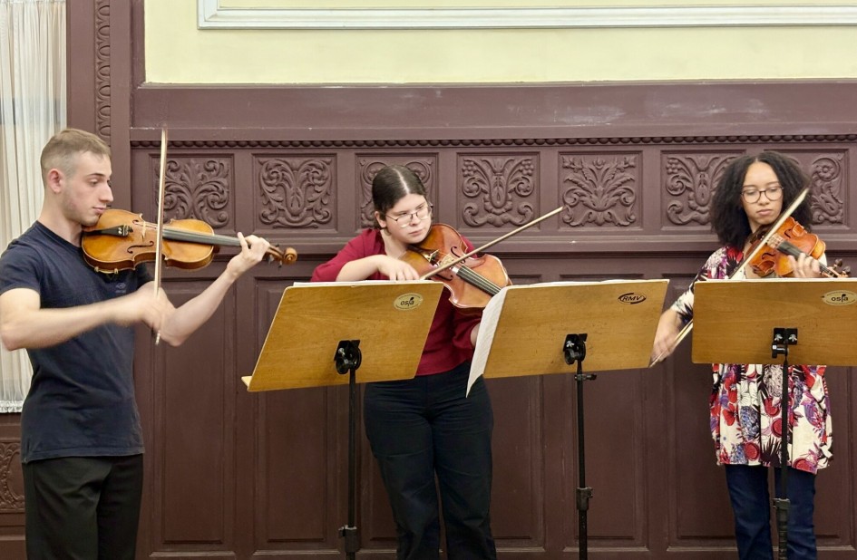 Trio de jovens violinistas em performance musical no saguão do prédio. Há pedestais com partituras diante dos instrumentistas