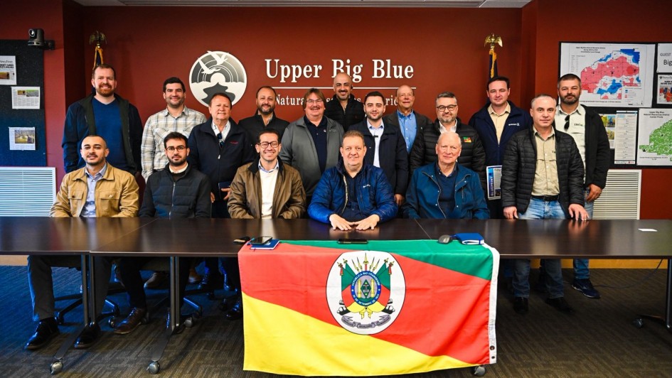 Grupo de 17 homens posa para foto em uma sala de reuniões com parede avermelhada, onde se lê “Upper Big Blue Natural Resources District”. À frente da mesa, uma bandeira do Rio Grande do Sul está estendida. O vice-governador Gabriel Souza aparece ao centro, sentado, com jaqueta azul escura.