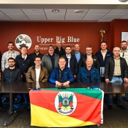 Grupo de 17 homens posa para foto em uma sala de reuniões com parede avermelhada, onde se lê “Upper Big Blue Natural Resources District”. À frente da mesa, uma bandeira do Rio Grande do Sul está estendida. O vice-governador Gabriel Souza aparece ao centro, sentado, com jaqueta azul escura.