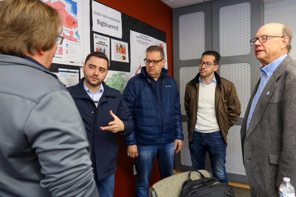 O vice-governador Gabriel Souza (ao centro, de jaqueta azul e camisa clara) conversa com quatro homens em um ambiente interno. Atrás deles há mapas e painéis informativos fixados na parede. Ele gesticula enquanto fala, em clima de atenção e diálogo.