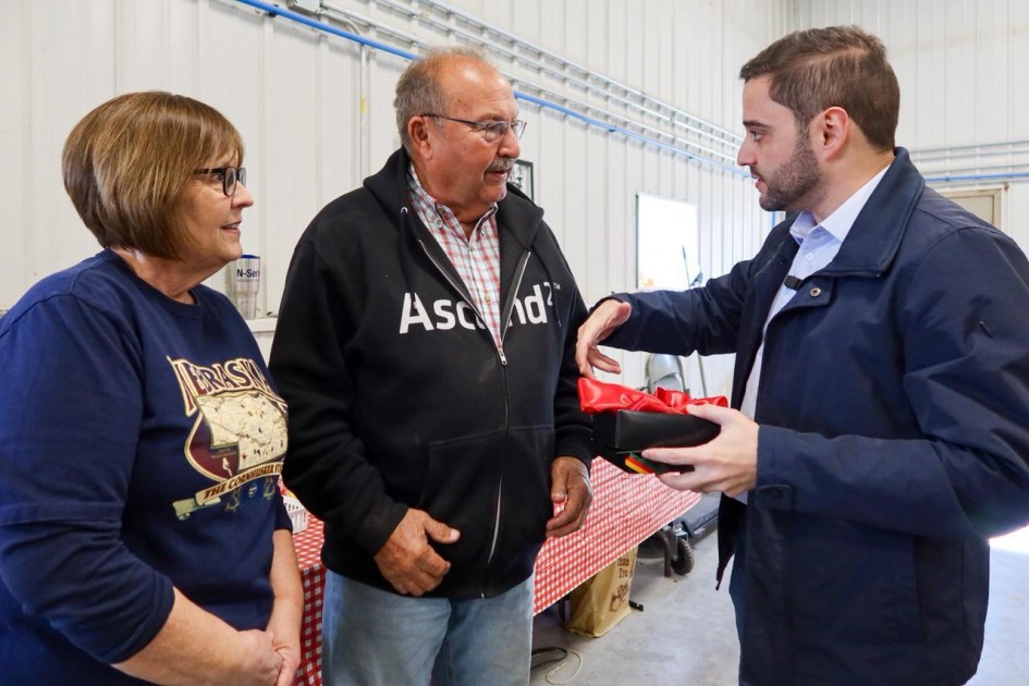 O vice-governador Gabriel Souza (à direita) conversa com um casal de agricultores. Eles estão sorridentes em um espaço interno com mesa coberta por toalha xadrez vermelha e branca.