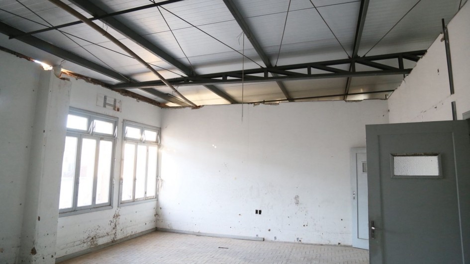 Foto mostra o pavimento, com uma sala vazia, de paredes brancas com duas janelas e uma porta cinza, onde aparece o telhado já reformado. 