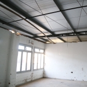 Foto mostra o pavimento, com uma sala vazia, de paredes brancas com duas janelas e uma porta cinza, onde aparece o telhado já reformado. 