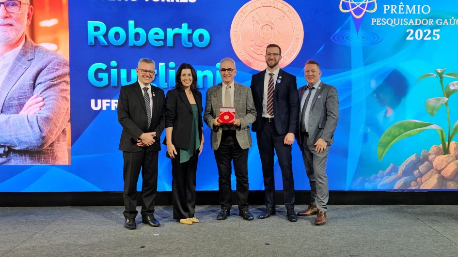 Cinco pessoas posam sorridentes diante do painel azul do Prêmio Pesquisador Gaúcho 2025. Ao centro, o pesquisador Roberto Giugliani, da UFRGS, segura o troféu em destaque, cercado por representantes do governo e da comunidade científica.