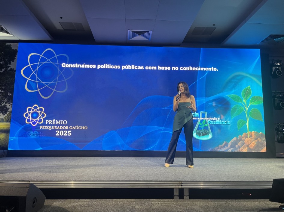 Mulher apresenta o evento no palco diante de um grande painel digital com a frase “Construímos políticas públicas com base no conhecimento”. O fundo exibe o logotipo do Prêmio Pesquisador Gaúcho 2025 e ilustrações de um frasco de laboratório e uma planta, simbolizando ciência e sustentabilidade.