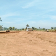 A foto mostra um terreno amplo de solo avermelhado. A área está limpa, sem vegetação, com marcas de máquinas no chão. Ao fundo, há alguns tratores e caminhões posicionados próximos a montes de terra.