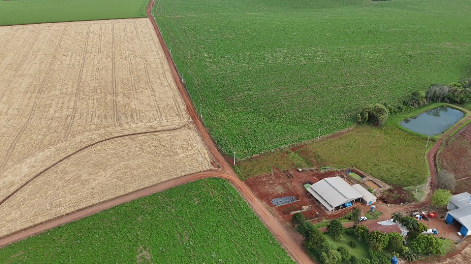 A foto aérea mostra uma área rural com grandes lavouras divididas em seções e uma propriedade rural com casa e galpões.