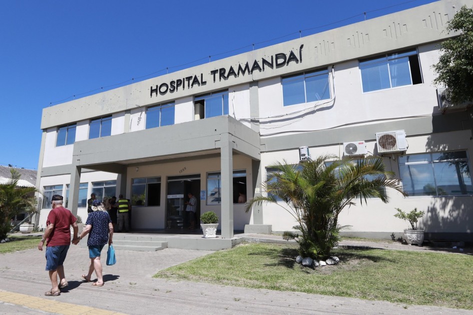 Fachada do Hospital Tramandaí, em um dia ensolarado. O prédio é claro, com janelas amplas e letreiro preto no topo. Em primeiro plano, um casal de idosos caminha de mãos dadas em direção à entrada principal.