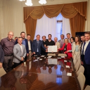 Uma foto em ambiente interno que captura um grupo de cerca de 16 pessoas (homens e mulheres) em pé, posando ao redor de uma grande e lustrosa mesa de reunião de madeira escura. O homem no centro da foto (de camisa preta) e a mulher ao lado dele (de blazer vermelho) estão segurando um documento estendido, possivelmente um termo de regulamentação ou lei, e todos estão olhando para a câmera. Os demais indivíduos vestem trajes sociais ou casuais de negócios, como ternos, blazers e camisas sociais.

O ambiente é formal e opulento, com paredes claras, cortinas de veludo marrom emoldurando uma janela ao fundo, um lustre no teto e objetos de decoração (incluindo faixas ou estandartes coloridos) no canto esquerdo. A sala está bem iluminada. O grupo parece ser formado por autoridades e membros do governo envolvidos na regulamentação de um programa de saúde.
