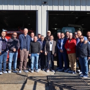 Uma foto externa de um grupo de cerca de 18 homens posando na frente da entrada aberta de um galpão. O grupo, vestindo roupas casuais de negócios ou casuais (muitos de jeans e blazers ou casacos), parece ser uma comitiva em visita. Todos usam protetores de sapatos (propés) de plástico azul-claro ou branco. Atrás deles, dentro do galpão, são visíveis grandes equipamentos agrícolas, incluindo uma máquina vermelha à esquerda e um equipamento verde escuro com a palavra "GENERAL" no centro. O galpão tem portas de correr de metal cinza. O grupo está em uma área de pátio com cercas de metal marrom de manejo de gado em primeiro plano, sugerindo que a visita é a uma instalação de produção animal ou agrícola de grande escala. O dia é de sol, com sombras projetadas no chão.