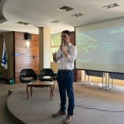 Uma foto em um auditório ou sala de reuniões. Um homem, de pé no centro, está falando em um microfone, gesticulando com as mãos abertas. Ele veste uma camisa social branca de manga comprida, jeans escuros e sapatos casuais marrons. Atrás dele, à direita, há uma tela de projeção exibindo um slide com o título "PROJETO DE MODERNIZAÇÃO Porto de Porto Alegre" e o logo da "Portos RS". À esquerda do homem, há duas poltronas pretas com braços e uma pequena mesa baixa de madeira entre elas. Mais à esquerda, em frente a uma parede de madeira escura, há três mastros com bandeiras: a do Rio Grande do Sul (amarela, vermelha e verde), outra azul e branca (possivelmente de uma universidade) e a do Brasil. O ambiente tem iluminação clara, e o piso é um carpete cinza em formato de semicírculo ou degraus. O homem está apresentando propostas em um evento chamado "Porto em Movimento".