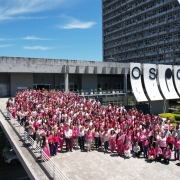 Uma foto aérea de um grande grupo de pessoas, predominantemente mulheres, vestidas em tons de rosa e branco, reunidas em uma esplanada ao ar livre em frente a um prédio. A foto foi tirada para marcar o "Outubro Rosa". O grupo está posicionado na rampa e na área pavimentada em frente a um edifício cinza de arquitetura moderna, com um letreiro que parece dizer "CAFF". Ao fundo e à direita, eleva-se um arranha-céu branco e curvo. O dia está ensolarado com céu azul. A legenda original da foto menciona "Servidores estaduais prestigiam foto coletiva do Outubro Rosa no CAFF" (Servidores públicos estaduais prestigiam foto coletiva do Outubro Rosa no CAFF).
