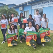 Foto posada de 11 estudantes adolescentes uniformizados em frente à escola, num gramado. Eles exibem cartazes de divulgação do Saeb.