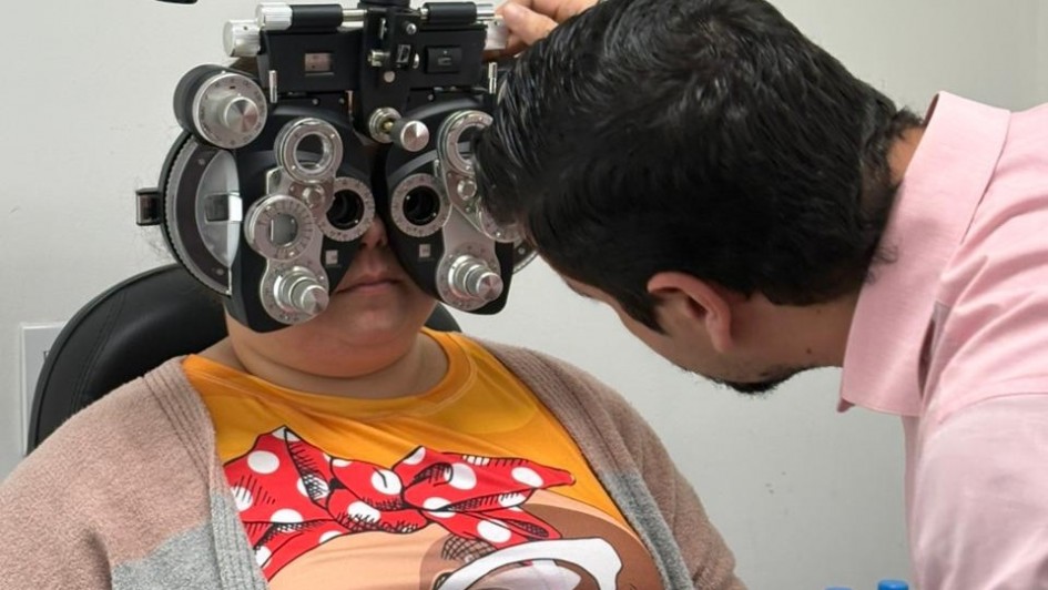 Pessoa fazendo exame de vista com aparelho de lentes, acompanhado por profissional. Estão em ambiente clínico, com garrafas d'água próximas.