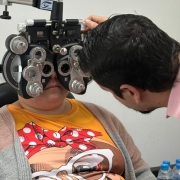 Pessoa fazendo exame de vista com aparelho de lentes, acompanhado por profissional. Estão em ambiente clínico, com garrafas d'água próximas.