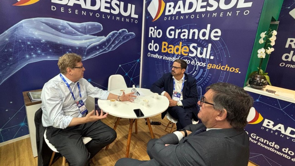 A imagem mostra três homens sentados ao redor de uma mesa redonda, conversando entre si. Ao fundo, há cartazes com a inscrição "Badesul Desenvolvimento" e "Rio Grande do Badesul".