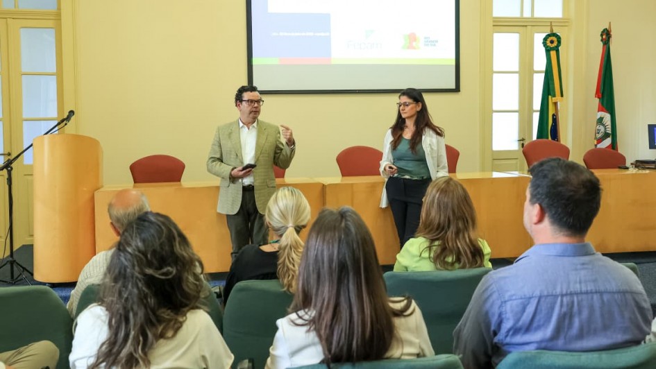Uma foto em um ambiente interno de um evento ou palestra, provavelmente pela manhã, como sugere o título do arquivo.

Em primeiro plano, a plateia está sentada em cadeiras escuras, com foco nas costas e na parte de trás da cabeça das pessoas, que estão assistindo à apresentação. O público é composto por homens e mulheres.

Ao fundo, em pé, em frente a uma mesa comprida de cor bege com detalhes em laranja, dois palestrantes estão falando. O homem à esquerda veste um blazer bege claro e segura o que parece ser um celular ou controle remoto. A mulher à direita veste uma blusa verde-água e um blazer branco.

Acima deles, uma tela de projeção exibe um slide com texto, a palavra "Palestra:" visível, e logotipos de organizações, incluindo "LABI" e "Fepam". O ambiente é formal, com paredes claras, grandes janelas à esquerda e à direita, e duas bandeiras em mastros à direita da tela, uma delas do Estado do Rio Grande do Sul.