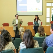 Uma foto em um ambiente interno de um evento ou palestra, provavelmente pela manhã, como sugere o título do arquivo.

Em primeiro plano, a plateia está sentada em cadeiras escuras, com foco nas costas e na parte de trás da cabeça das pessoas, que estão assistindo à apresentação. O público é composto por homens e mulheres.

Ao fundo, em pé, em frente a uma mesa comprida de cor bege com detalhes em laranja, dois palestrantes estão falando. O homem à esquerda veste um blazer bege claro e segura o que parece ser um celular ou controle remoto. A mulher à direita veste uma blusa verde-água e um blazer branco.

Acima deles, uma tela de projeção exibe um slide com texto, a palavra "Palestra:" visível, e logotipos de organizações, incluindo "LABI" e "Fepam". O ambiente é formal, com paredes claras, grandes janelas à esquerda e à direita, e duas bandeiras em mastros à direita da tela, uma delas do Estado do Rio Grande do Sul.