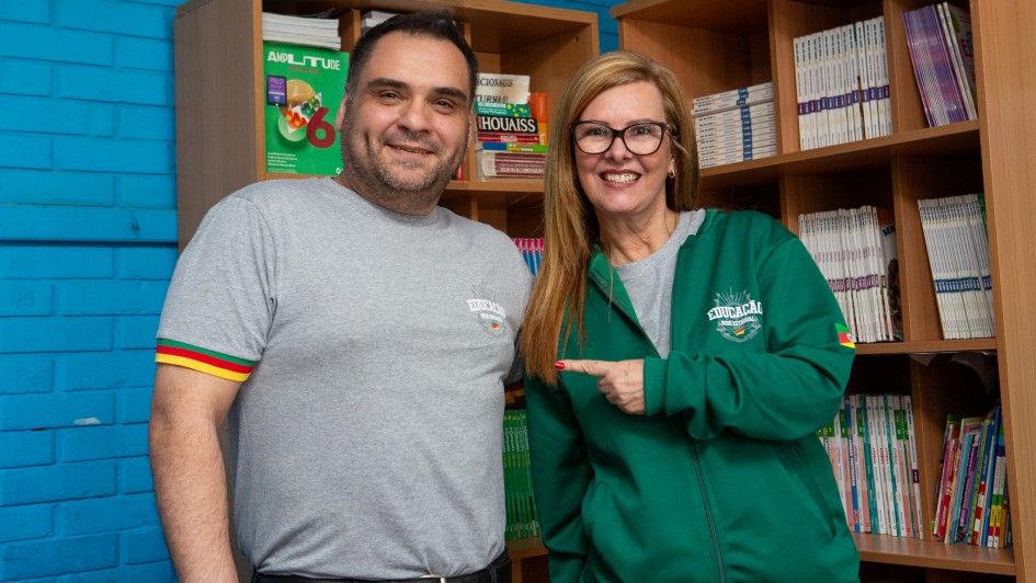 Um homem e uma mulher posam sorrindo diante de estantes com livros e materiais didáticos. Ambos vestem camisetas cinzas com o logotipo “Educação Rede Estadual”, e a mulher usa também uma jaqueta verde com o mesmo símbolo. Atrás deles, há prateleiras contendo exemplares de dicionários, livros didáticos e revistas escolares.