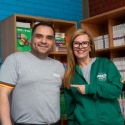 Um homem e uma mulher posam sorrindo diante de estantes com livros e materiais didáticos. Ambos vestem camisetas cinzas com o logotipo “Educação Rede Estadual”, e a mulher usa também uma jaqueta verde com o mesmo símbolo. Atrás deles, há prateleiras contendo exemplares de dicionários, livros didáticos e revistas escolares.