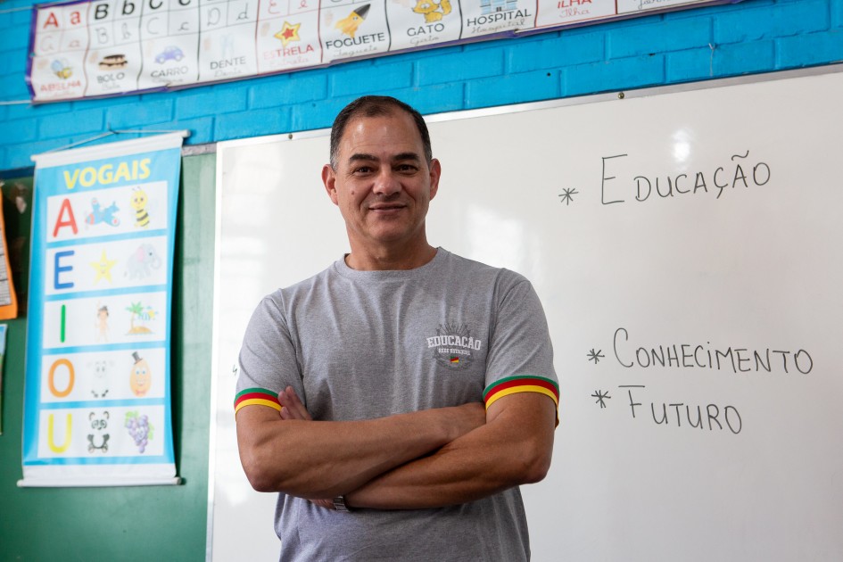 Um homem está em pé diante de um quadro branco, com os braços cruzados, vestindo camiseta cinza com o logotipo “Educação Rede Estadual”. No quadro, aparecem escritas as palavras “Educação”, “Conhecimento” e “Futuro”. Ao lado, há cartazes coloridos com ilustrações das vogais e imagens associadas a cada letra.