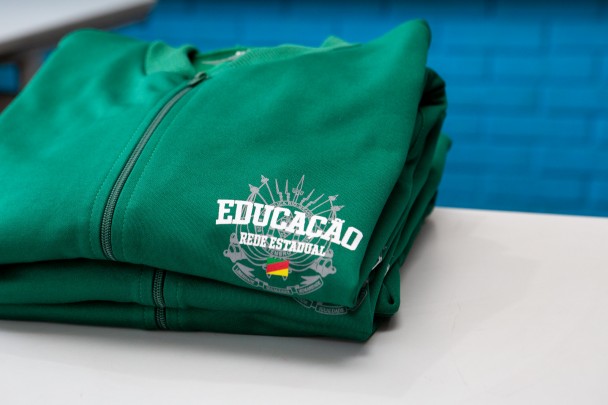 Um moletom verde dobrado está sobre uma mesa branca. Na parte visível da peça, está estampado o logotipo “Educação Rede Estadual”, com o brasão do Rio Grande do Sul abaixo.