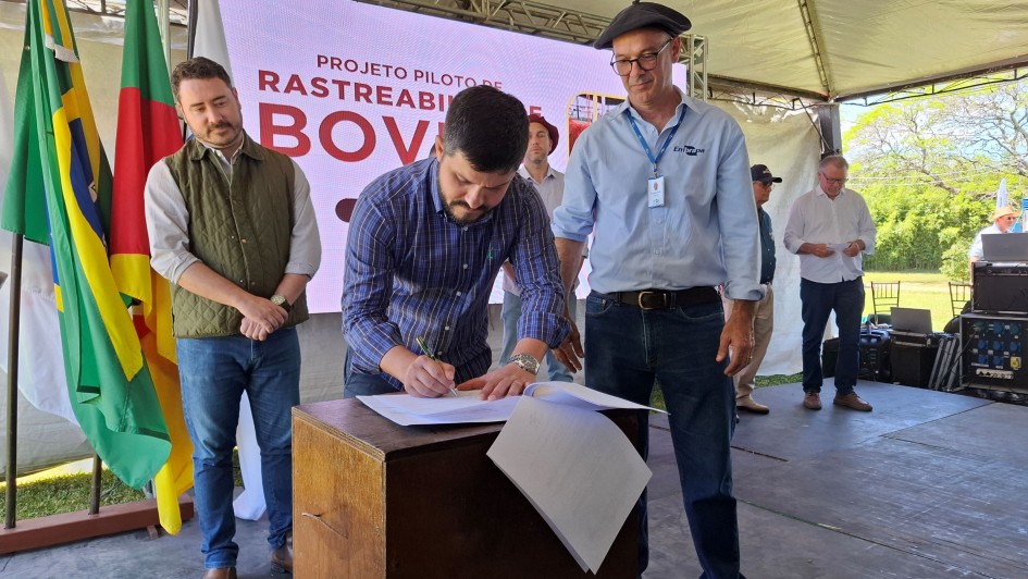 A imagem mostra um homem assinando um documento enquanto outros dois estão posicionados ao lado, olhando para ele. Ao fundo, há um painel indicando o tema do evento sobre rastreabilidade bovina.